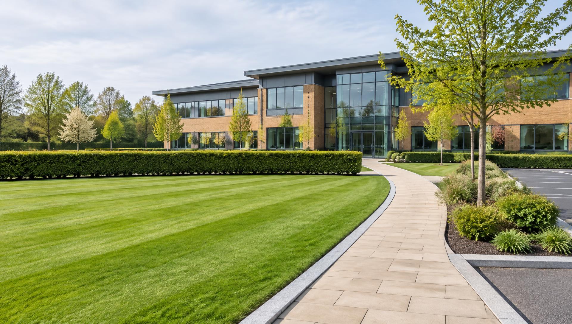 A modern UK business park with a striped mown lawn, clipped hedge, planted bed, paved footpath and an office building set back behind the grounds
